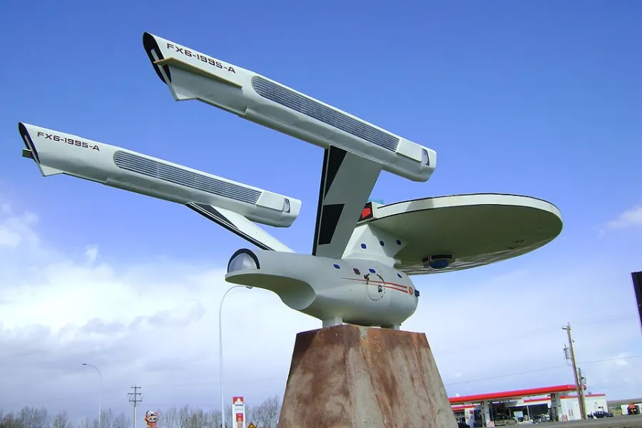 A white model of the starship Enterprise on a tall stone pedestal, seen from below against a blue sky, with FX6-1995-A painted on each of its two engines.
