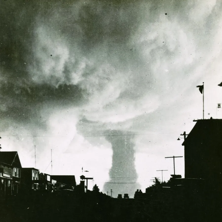 A dark street of low buildings and telegraph poles, with a tornado funnel reaching down from a pale, swirling sky at the end of it.