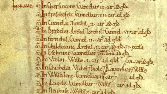 Lines of a Domesday Book folio in medieval Latin script, each beginning with a place name, with red strokes marking the entries.